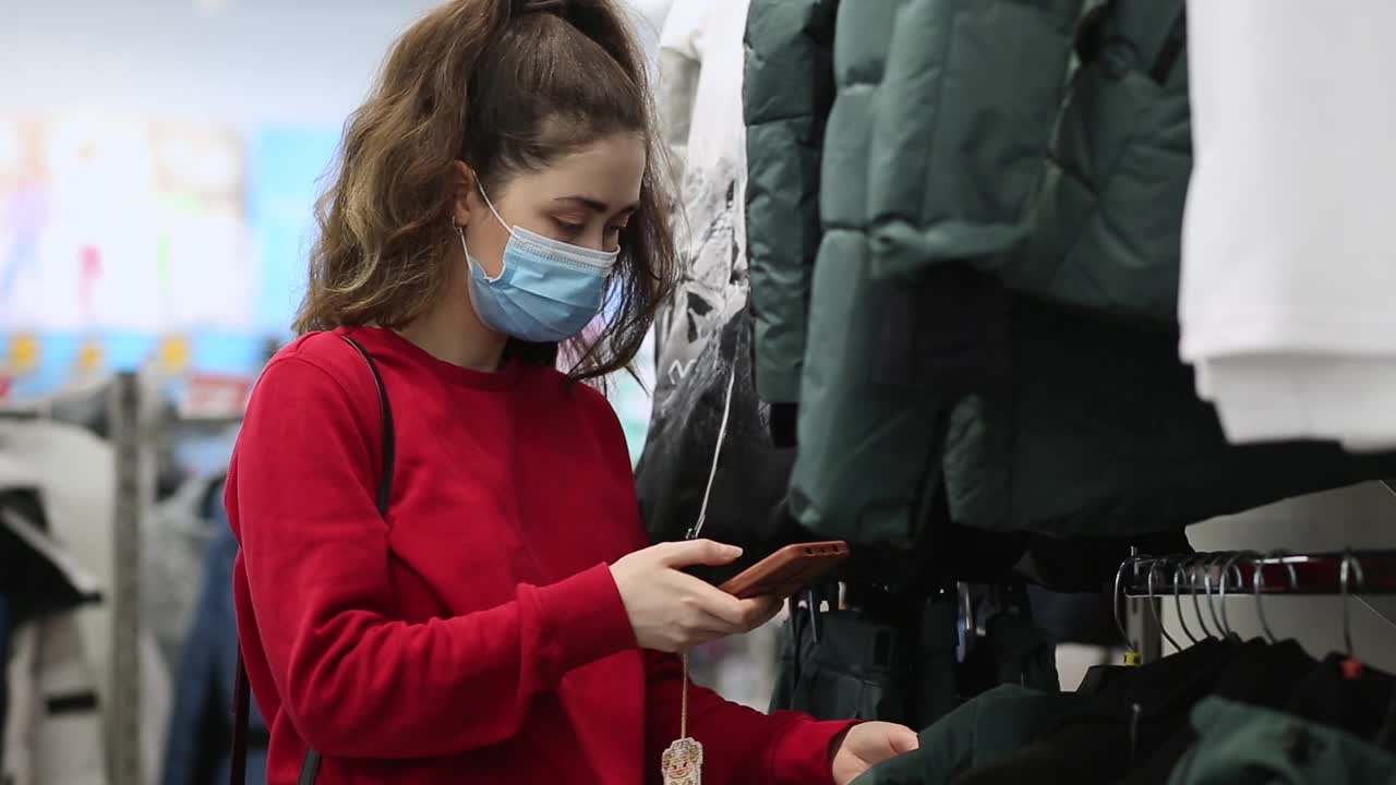 一名戴着医用口罩的年轻女子的肖像正在扫描衣服标签上的二维码。购物的概念和现代科技视频素材