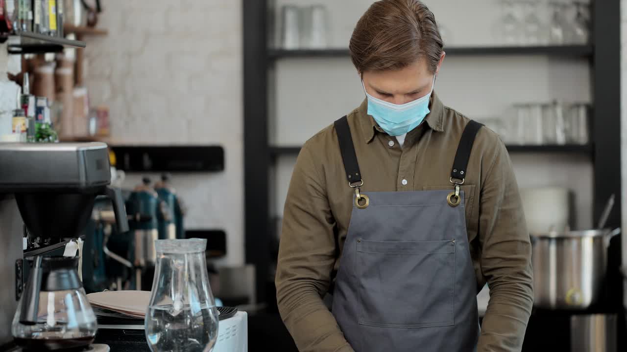 白人英俊的男性咖啡师的肖像，戴着医用口罩和手套，给相机一杯咖啡和微笑。在咖啡馆里，咖啡师端着咖啡外带。视频素材