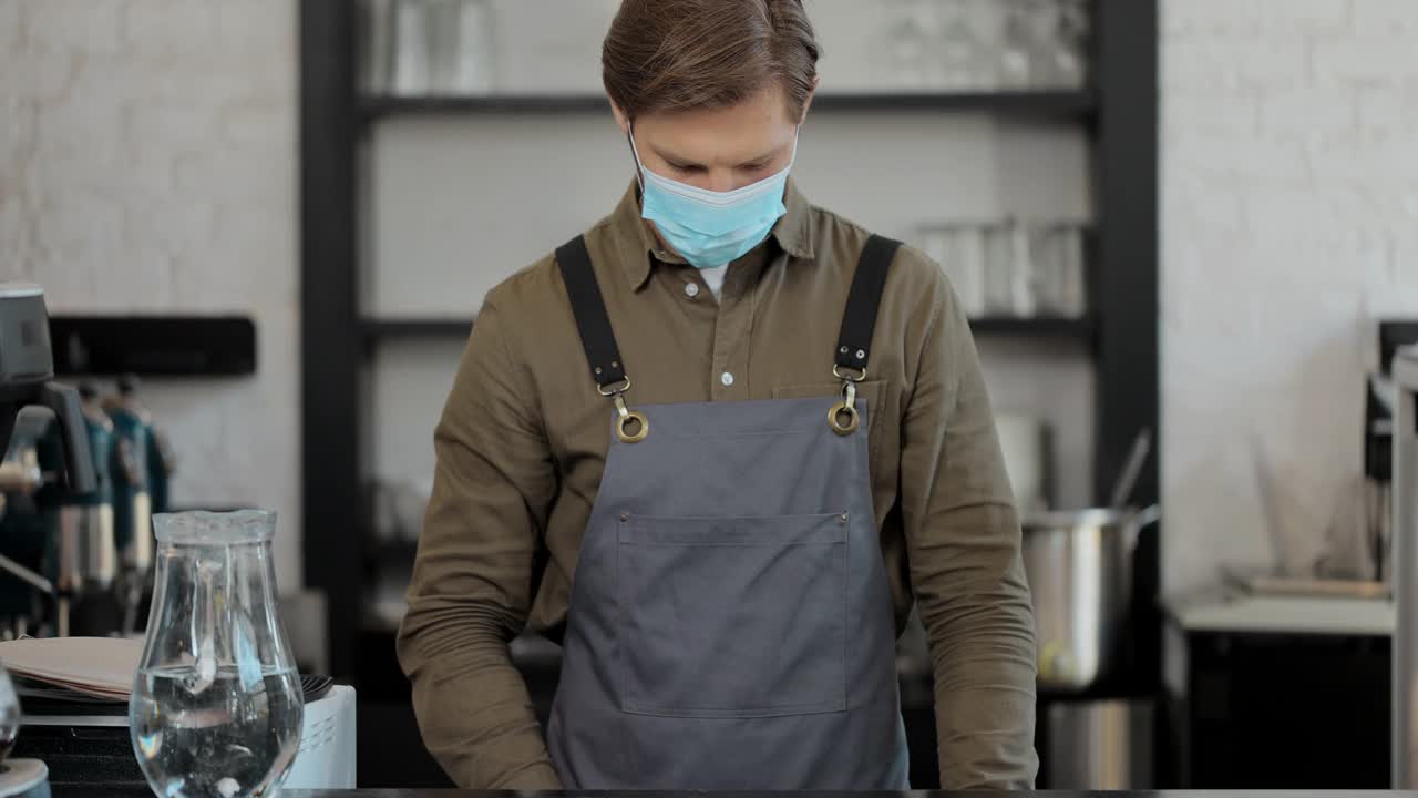 戴着医用口罩的男咖啡师的肖像，女服务员在城市咖啡馆拿着咖啡杯，心情愉快。咖啡店的概念。视频素材