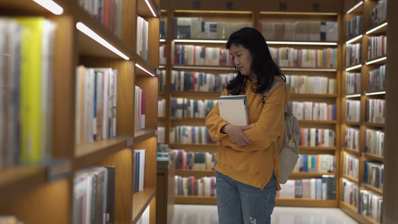 在图书馆，一位亚洲妇女在书架前走来走去，一边浏览书籍视频素材