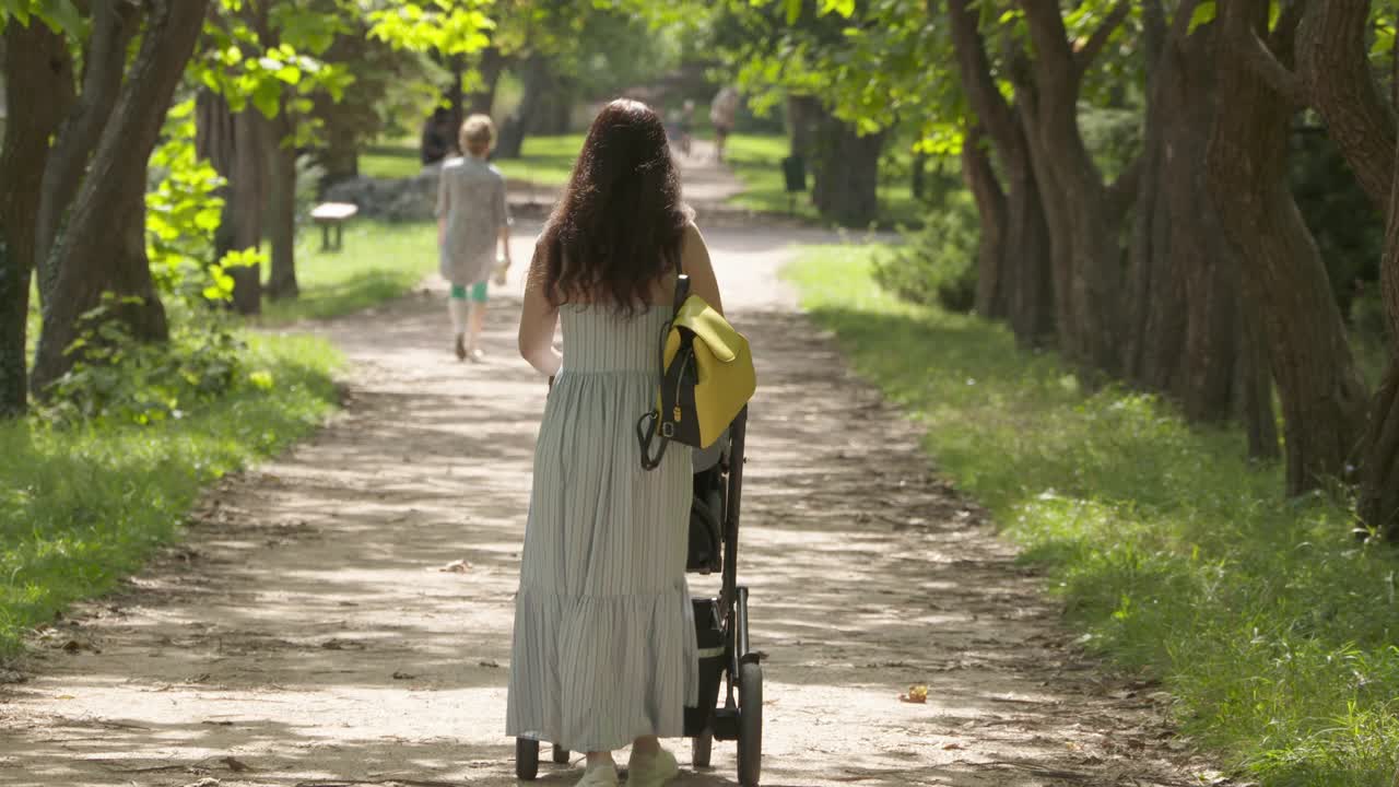 一个阳光明媚的夏日，妈妈推着婴儿车在公园小路上散步。视频素材