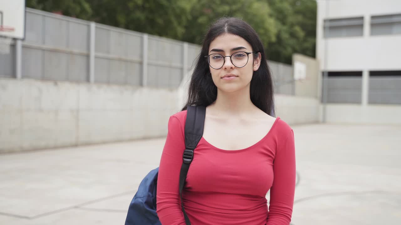 年轻的西班牙女大学生肖像黑发，眼镜和红色休闲服在中学的院子里视频素材