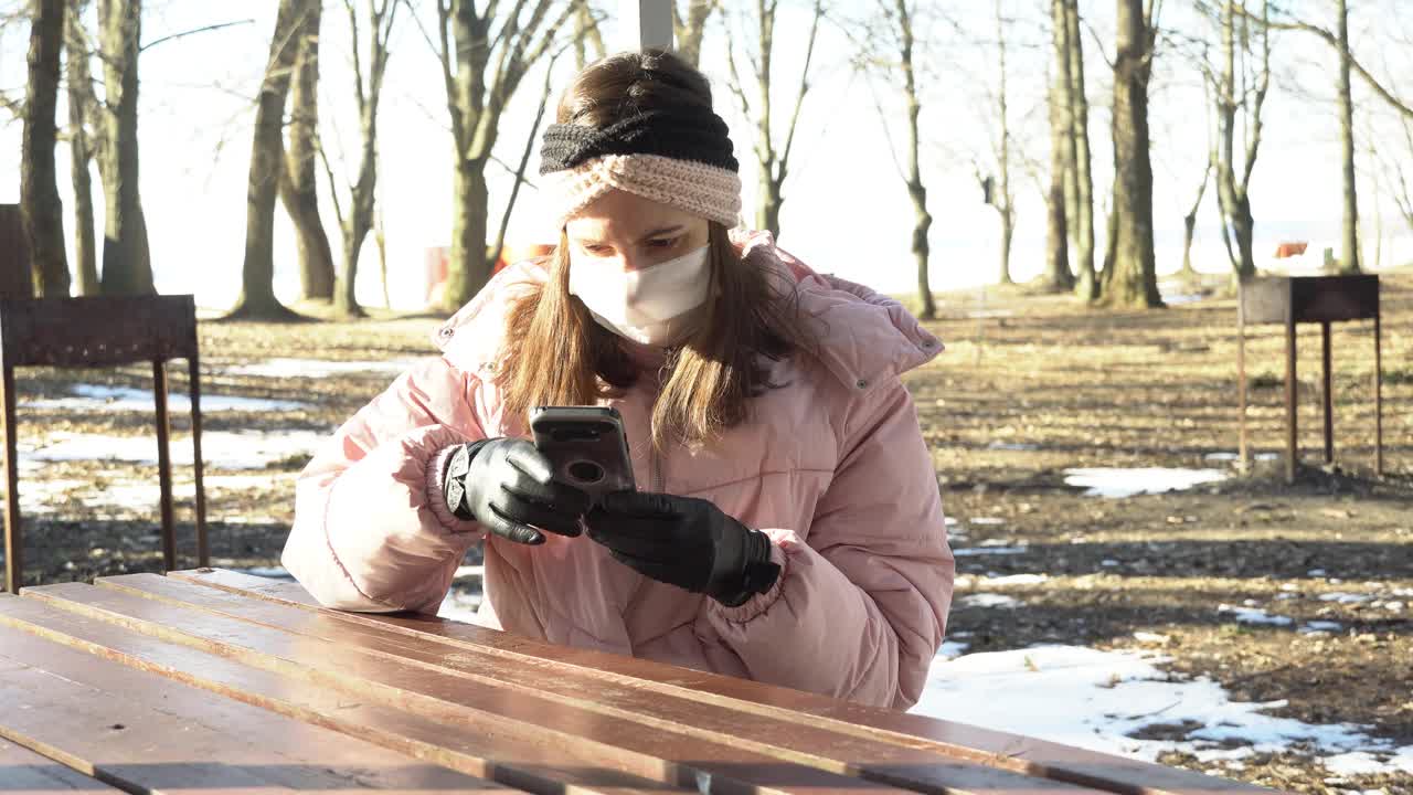 一名戴着防护口罩的女孩在冠状病毒期间在公园通过视频会议使用手机，并摘下手套使用该设备，这是冠状病毒限制措施视频素材