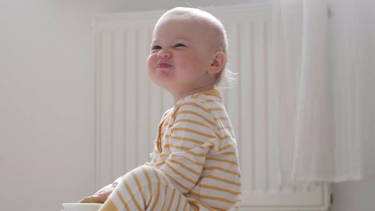 在家吃宝宝的照片。孩子做鬼脸。孩子坐在地板上，手里拿着碗。健康食品，午餐时间视频素材