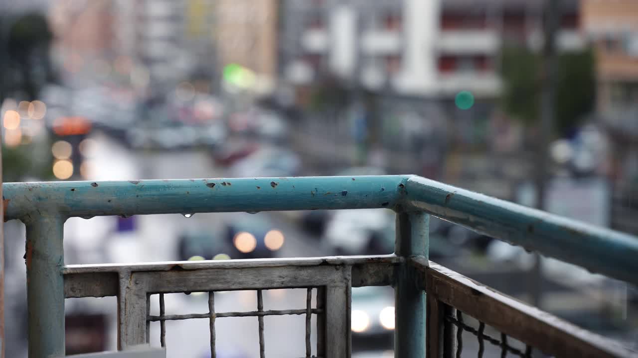 阴沉的雨天，从住宅楼阳台看城市景观，雨滴顺着陈旧生锈的阳台栏杆落下，流淌在模糊的城市街道交通背景上。街头生活视频素材