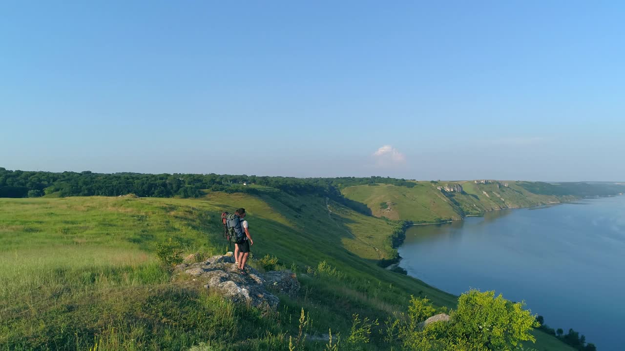 两名旅行者背着背包站在山顶上，俯瞰风景秀丽的河流视频素材