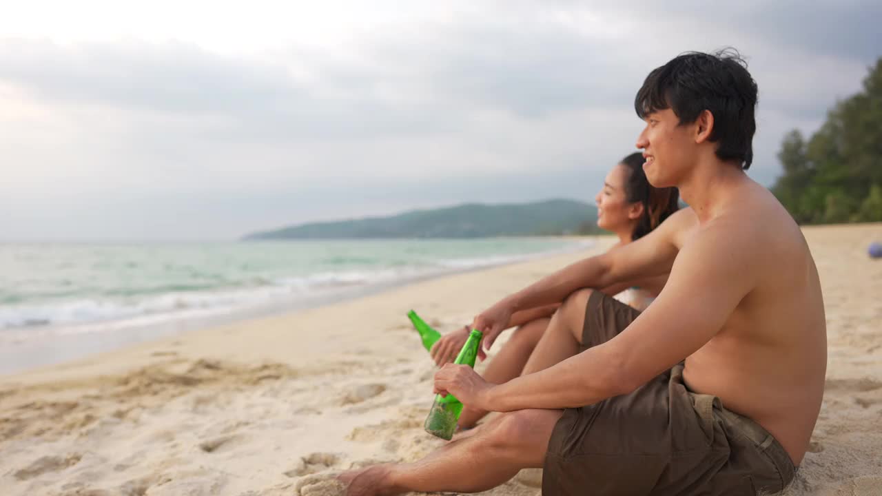 一群快乐的亚洲男女朋友坐在沙滩上，喝着啤酒，在夏日的夕阳下聊天。视频素材