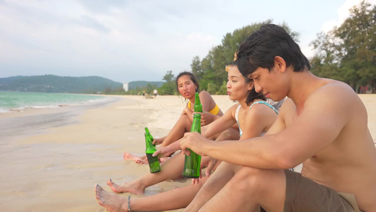 一群快乐的亚洲男女朋友坐在沙滩上，喝着啤酒，在夏日的夕阳下聊天。视频素材