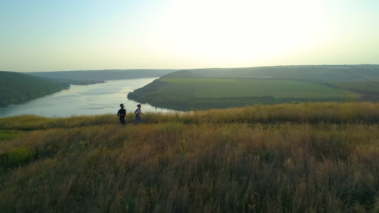 这对夫妇沿着绿色的小山慢跑，风景优美的河流为背景。慢动作视频素材