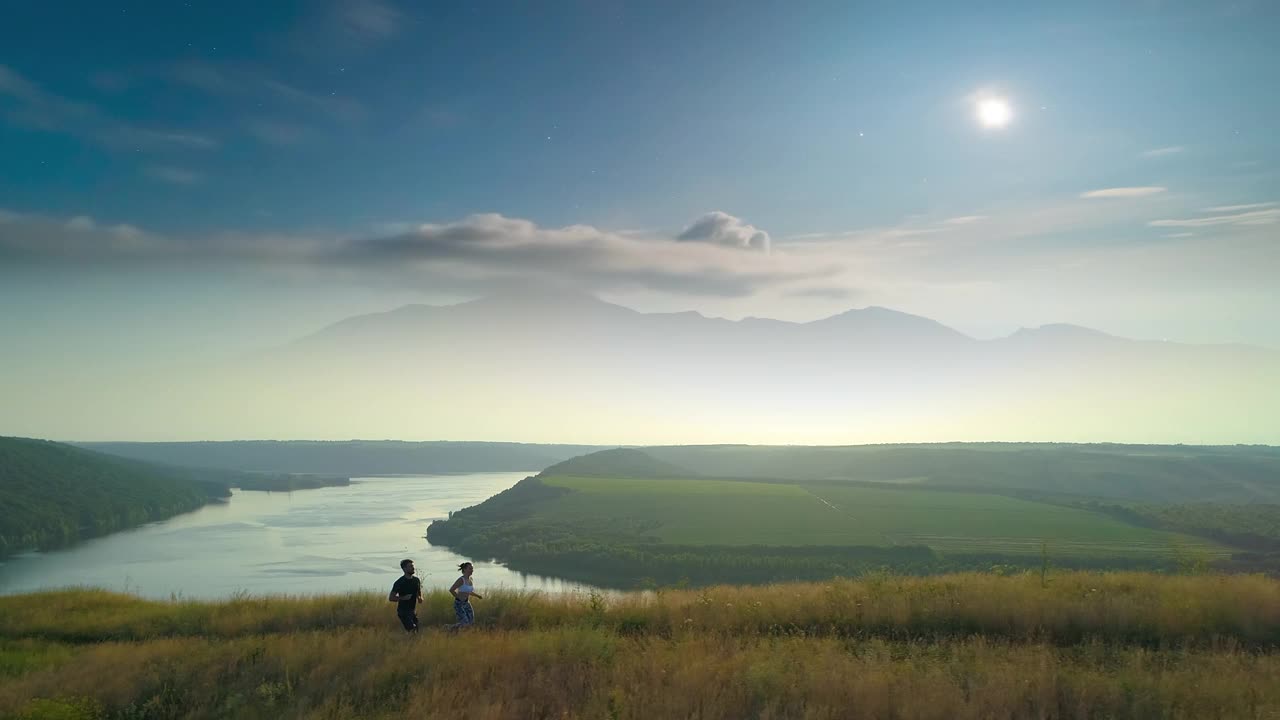 这对夫妇沿着绿色的小山慢跑，风景优美的河流为背景。慢动作视频素材