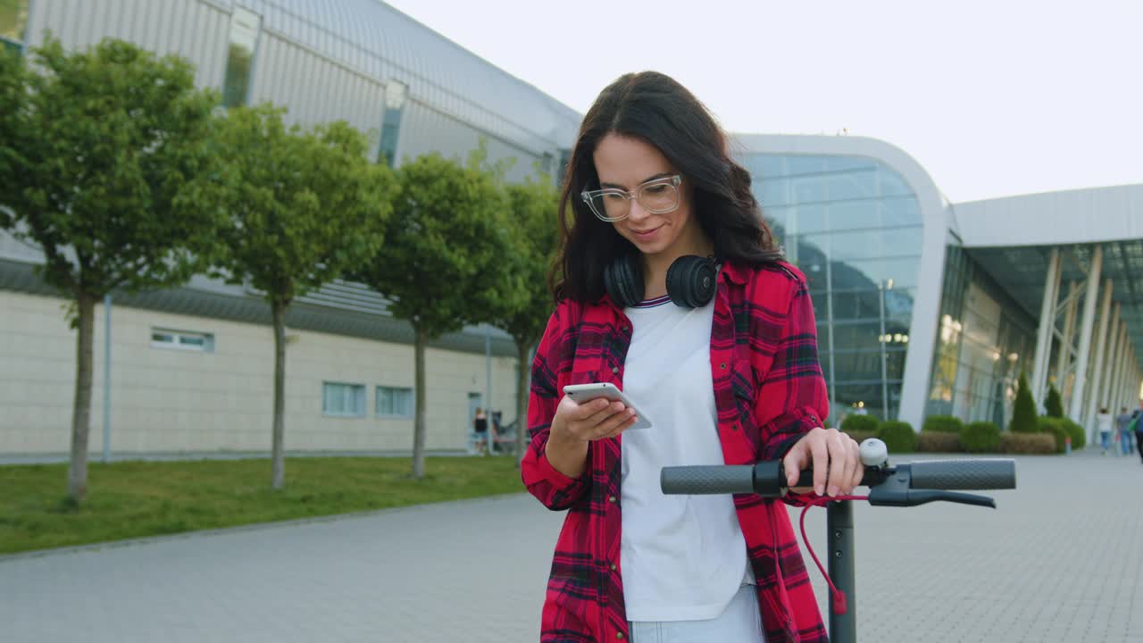 美丽的微笑满足了穿着休闲装的棕色头发的年轻时尚女孩，她骑着电动滑板车在有绿色植物的现代大建筑附近散步，并使用智能手机视频素材