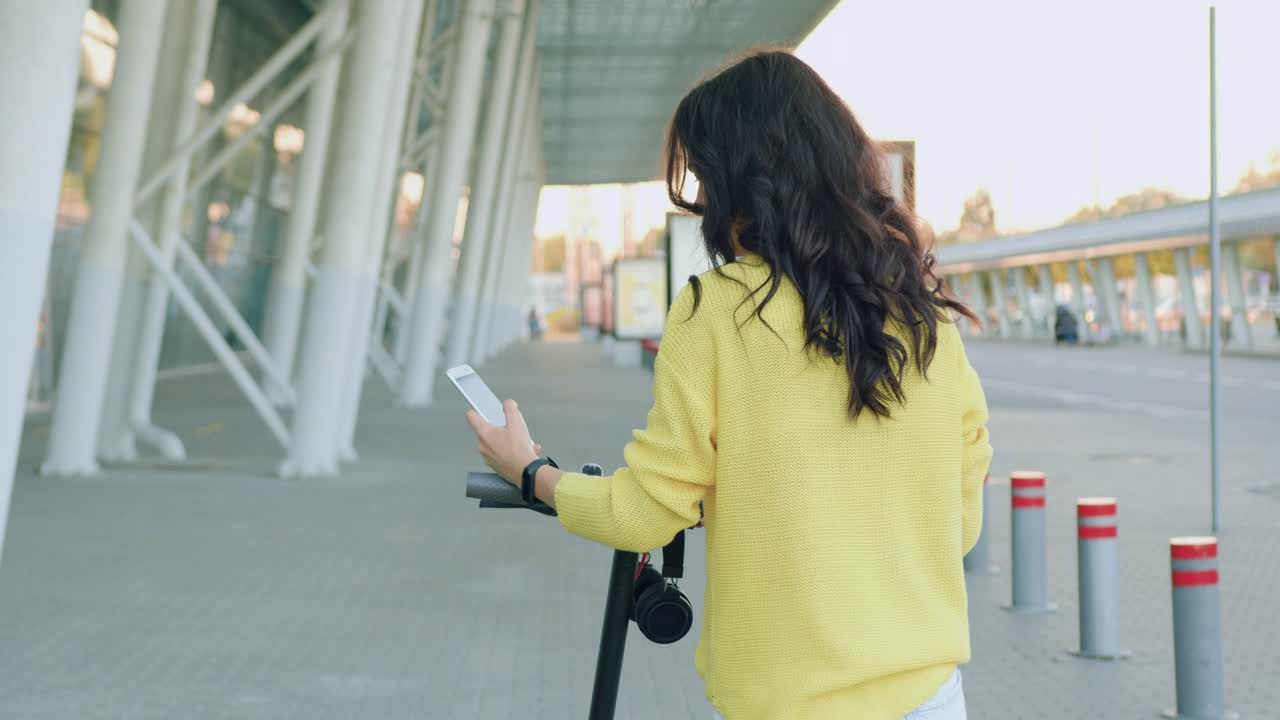 后视图，漂亮时尚的棕色头发的年轻女子，穿着时髦的衣服，走在电动滑板车和使用她的智能手机附近的大型城市建筑视频素材