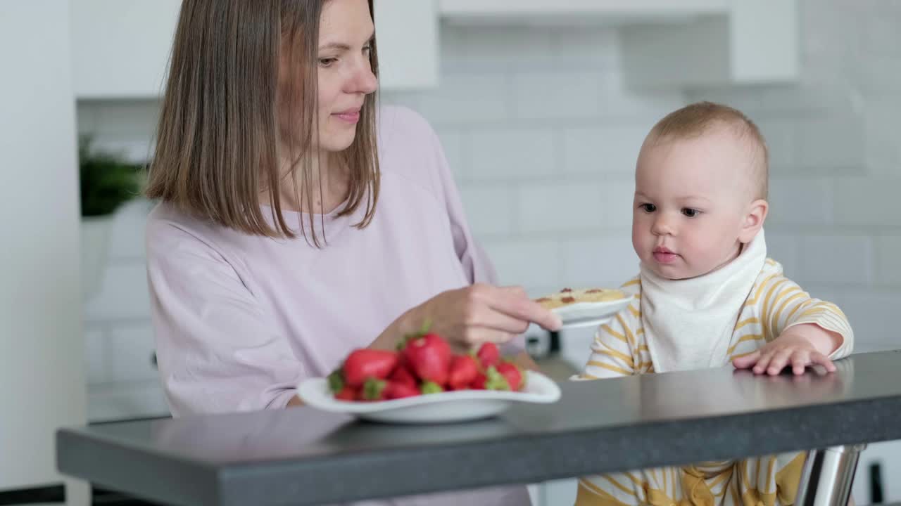 妈妈和宝宝在家里的厨房里做饭。健康食品，午餐时间。孩子孩子选择吃什么饼干还是草莓视频素材