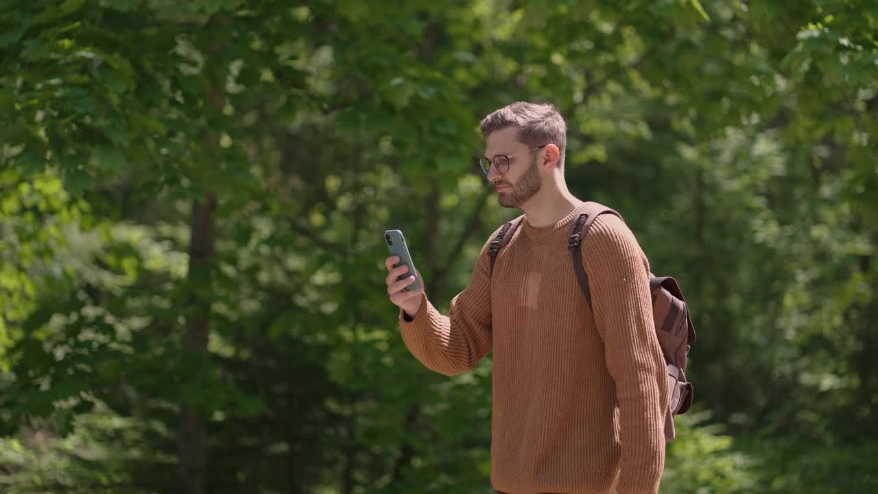 一名年轻的男性旅行者背着背包，带着手机在森林里行走，拍摄视频，拍摄自然风景。制作视频内容，在线播放。视频素材