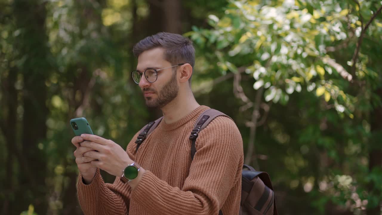 一名背着背包的男性旅行者在森林里的一条土路上行走，写信息，在互联网上交流，查看内容，在手机上做笔记，环顾四周。视频素材