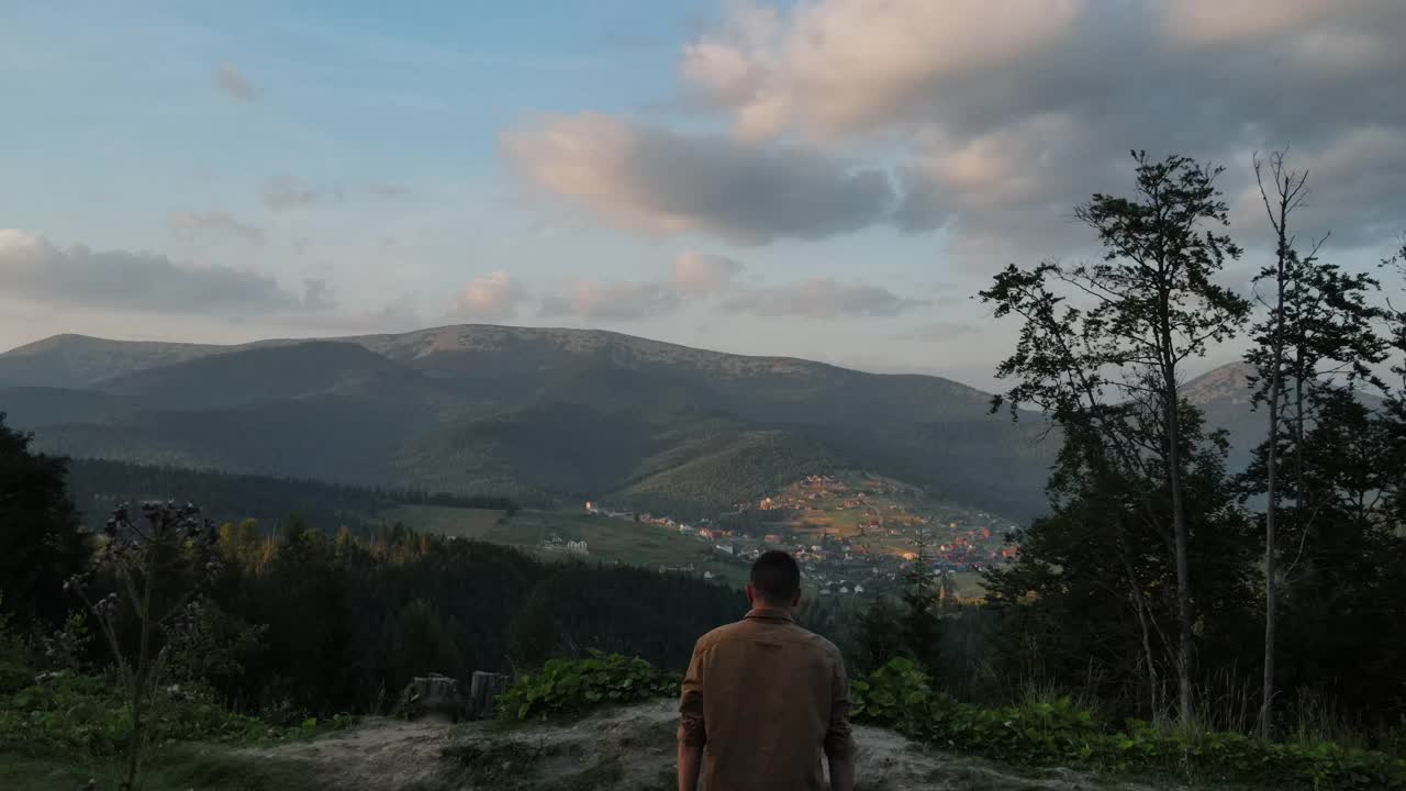 无人机跟随年轻男子在绿色的田野上奔跑在背景山的顶峰，在日落或日出时，举起双臂，快乐和醉心于生活，青春和幸福。旅游的概念视频素材