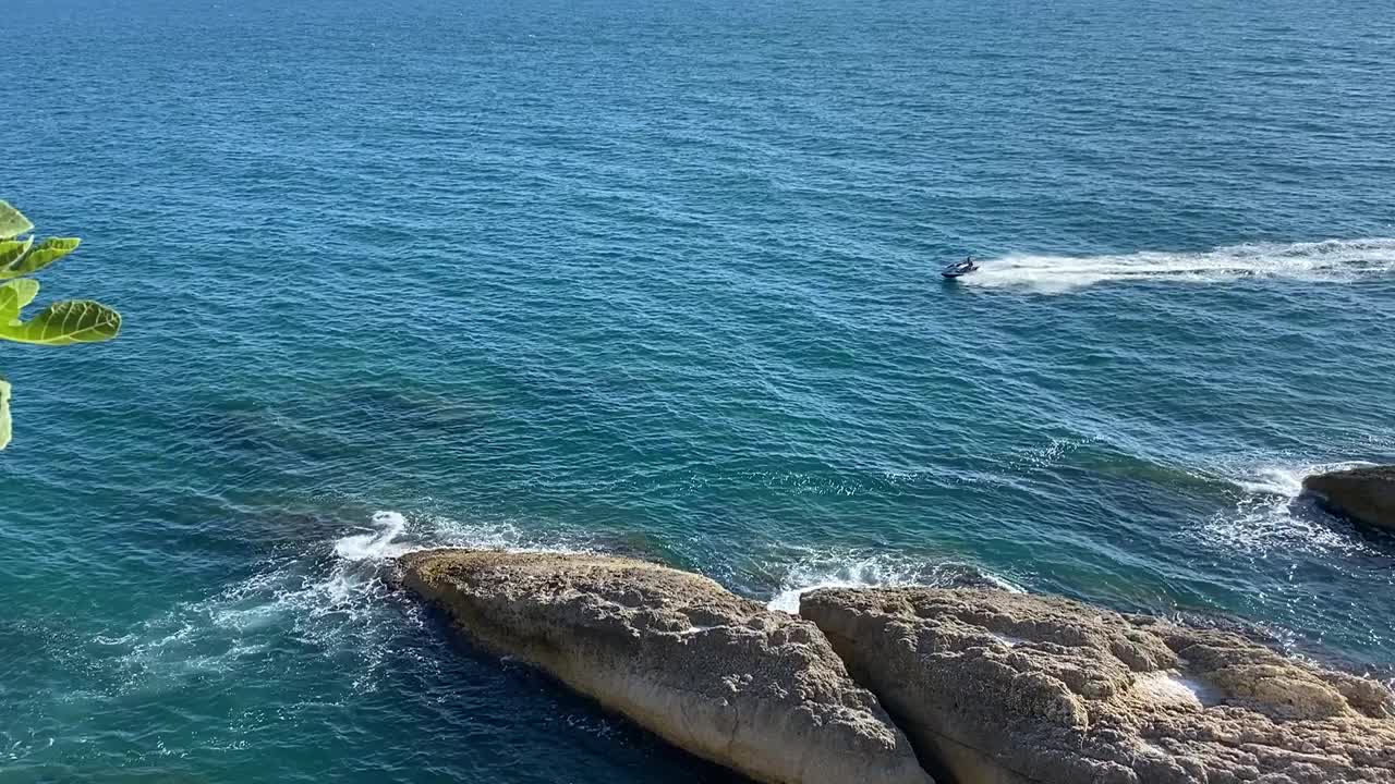 蓝色的海浪。摩托艇沿着多岩石的海岸快速地漂浮着。地中海夏日休闲。风景优美的海岸的岩石。美丽的海景。视频素材