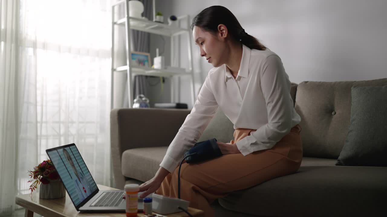 一位亚洲妇女病人在家里用笔记本电脑和她的医生聊天，同时使用血压检查视频素材