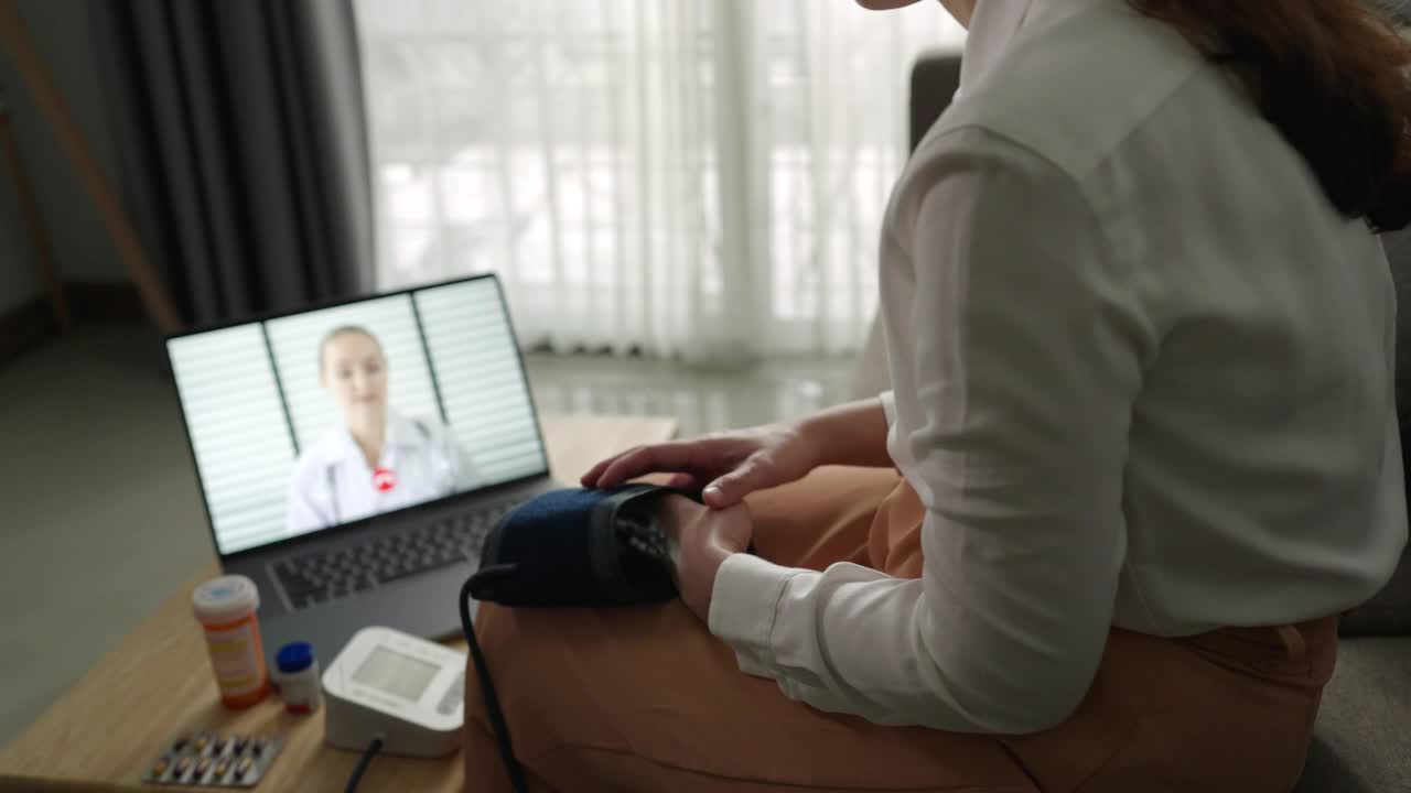 一位亚洲妇女病人在家里用笔记本电脑和她的医生聊天，同时使用血压检查视频素材