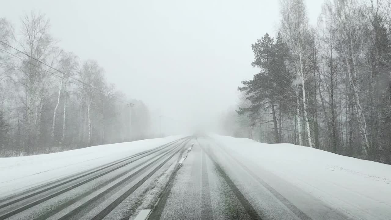 在冬天，在森林里，在大雪中能见度很低的情况下，透过汽车的挡风玻璃在一条空的、下雪的、结冰的道路上看视频素材