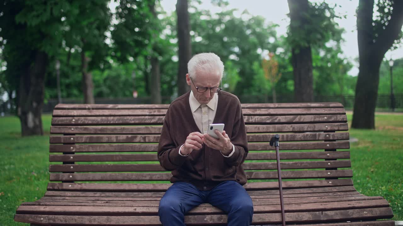 一名戴着眼镜的老年男子坐在长椅上，试图应付新的智能手机视频素材