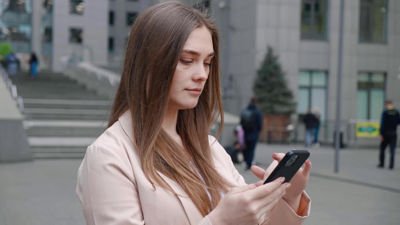 一名年轻女子站在办公楼外看着智能手机，震惊不已。带坏消息的令人不快的惊讶女性。视频素材