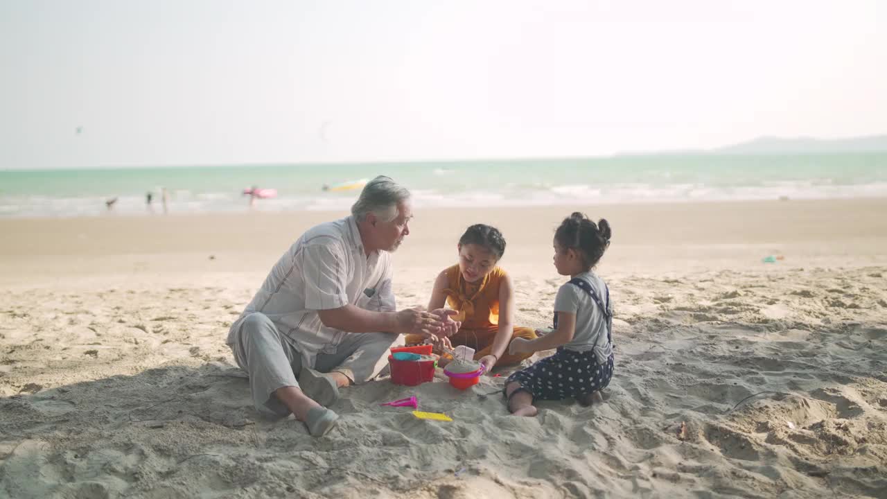 暑假期间，亚洲爷爷和两个孙女在海滩上玩沙滩玩具视频素材