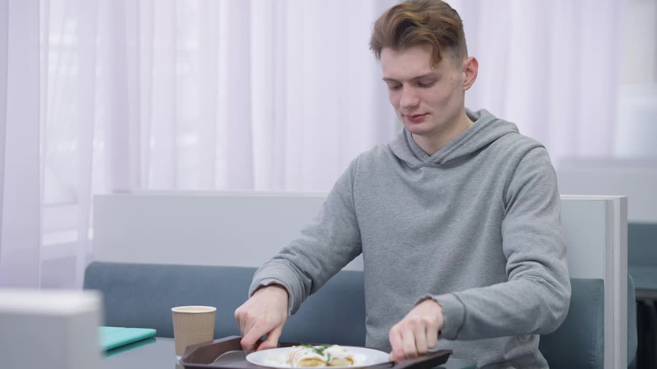 饥饿的年轻男学生在餐厅品尝美味的煎饼，脸上带着满意的表情。一名白人男子坐在餐厅里吃着美味的甜点。午休时间概念视频素材