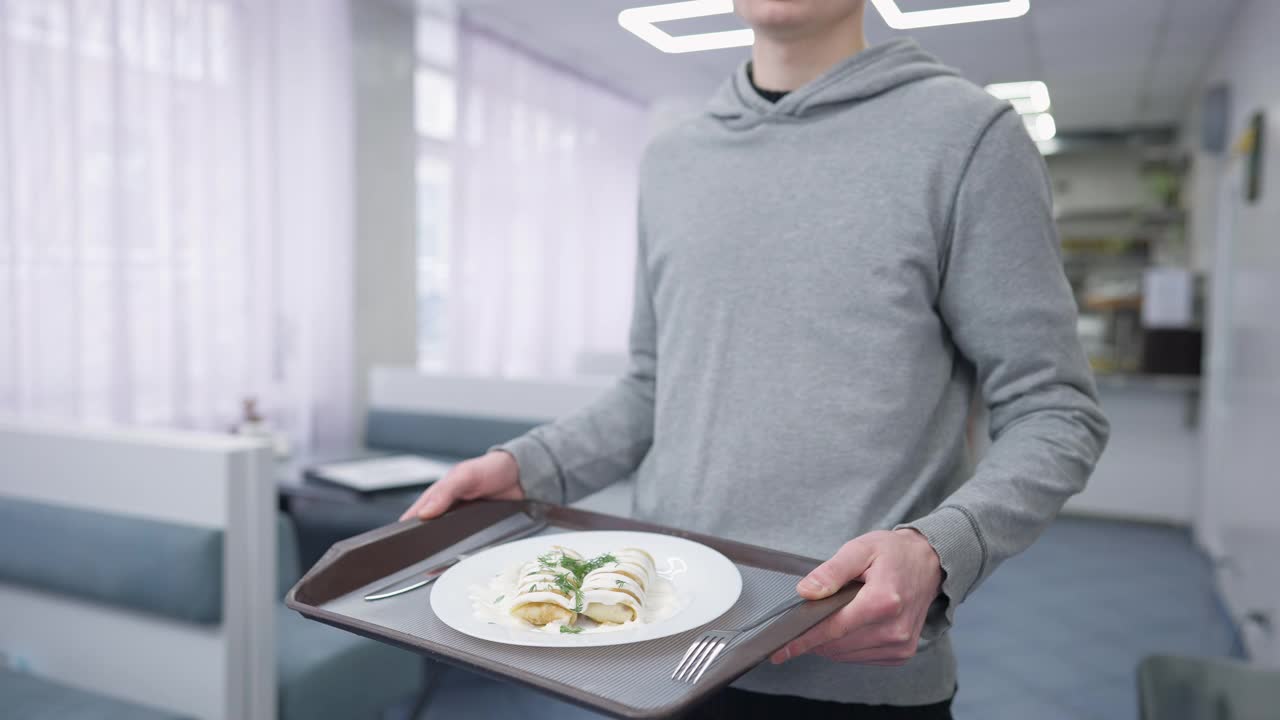 陌生的男学生拿着托盘和甜煎饼，叉子和餐刀站在室内自助餐厅。一名年轻的白人男子在食堂与美味的食物合影视频素材