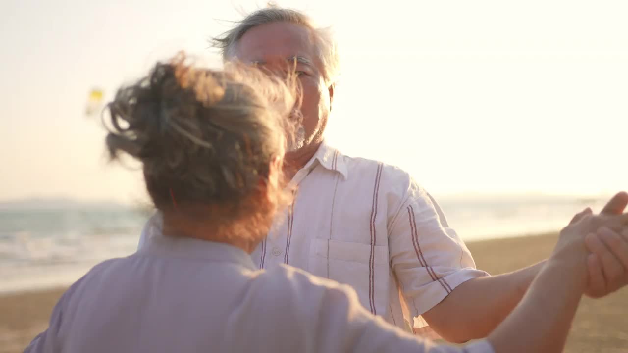 4K亚洲老年夫妇在夏日日落的海滩上享受浪漫的假期，翩翩起舞视频素材