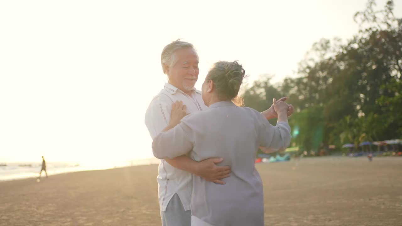 4K亚洲老年夫妇在夏日日落的海滩上享受浪漫的假期，翩翩起舞视频素材