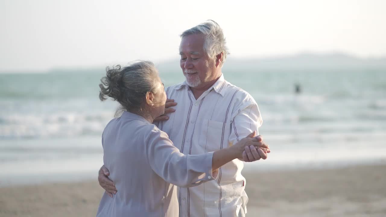 4K亚洲老年夫妇在夏日日落的海滩上享受浪漫的假期，翩翩起舞视频素材