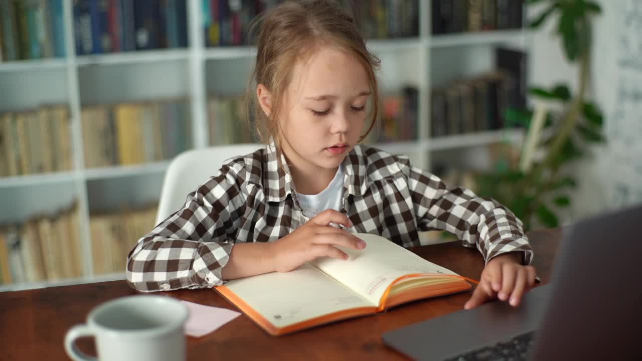 特写俯视图，聪明的小学女孩在做家庭作业，在笔记本上写笔记，看着笔记本电脑。视频素材