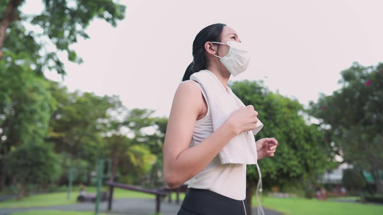 年轻健康的白种亚洲工人穿着运动服，脱下医用口罩，戴上耳机，在阳光明媚的日子里，在绿城公园享受着听音乐和锻炼身体的乐趣，健康的城市生活视频素材