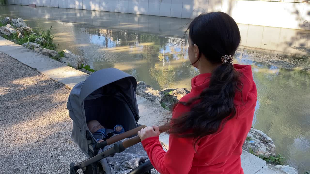 一个阳光明媚的夏日，妈妈推着婴儿车在公园小路上散步。视频素材