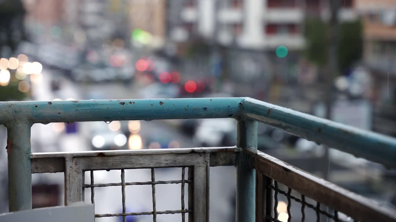 阴沉的雨天，从住宅楼阳台看城市景观，雨滴顺着陈旧生锈的阳台栏杆落下，流淌在模糊的城市街道交通背景上。街头生活视频素材