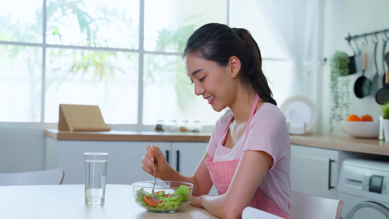 穿着围裙的亚洲美女在家里的厨房里吃着蔬菜沙拉。年轻美丽的女孩感到快乐，享受早餐与蔬菜健康的食物和清洁的水，以饮食的健康在家里。视频素材