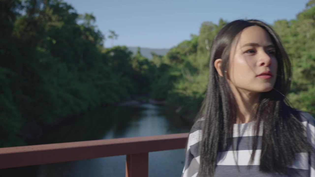 美丽的黑发游客在热带雨林徒步旅行时，独自走在木桥上，穿过小溪，阳光照在茂密的树叶上，女性的脸视频素材