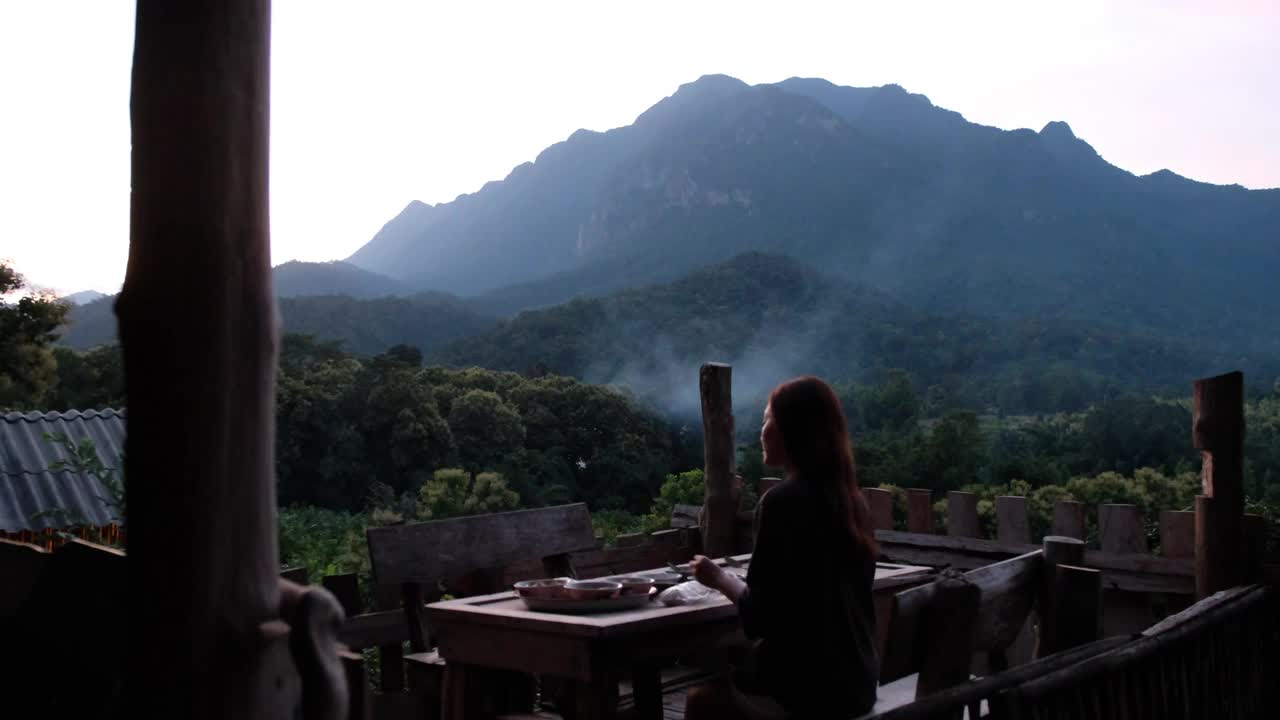 一个女人坐在阳台上吃晚饭，可以看到绿色的山景视频素材