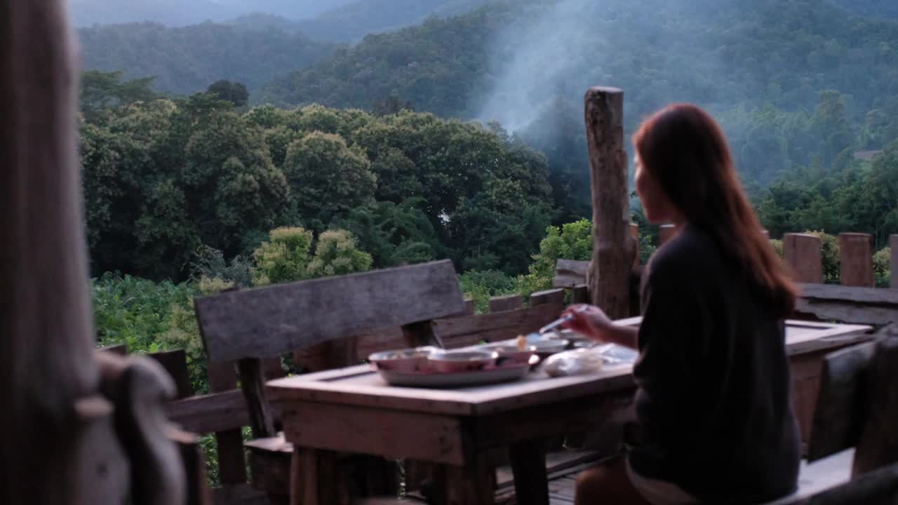 一个女人坐在阳台上吃晚饭，可以看到绿色的山景视频素材