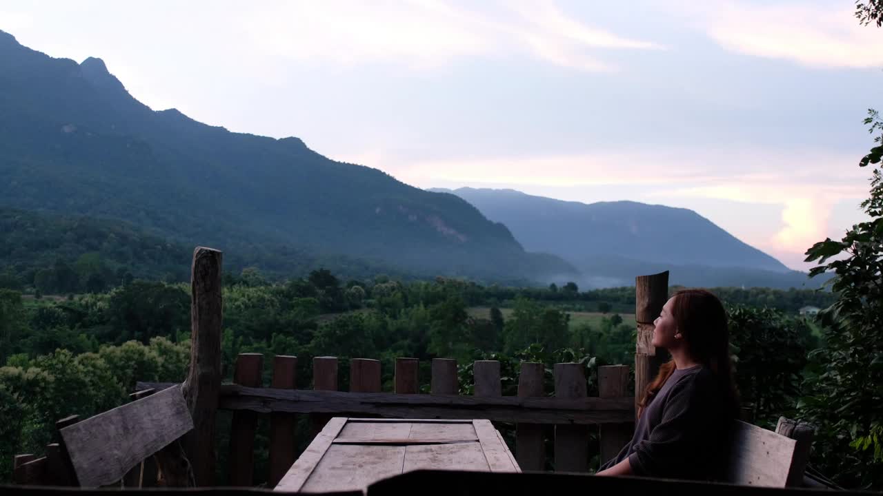 一位女游客坐在木制阳台上，看着美丽的山景视频素材