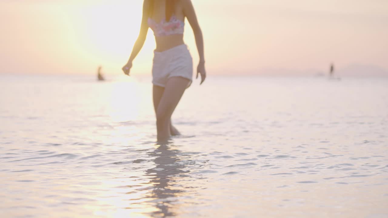 年轻女子在夏天的假期漫步在橙色金色阳光海岸的水，夏季海滩的目的地，浸泡在咸咸的海水，剪影女子放松，温暖的气候热带岛屿视频素材