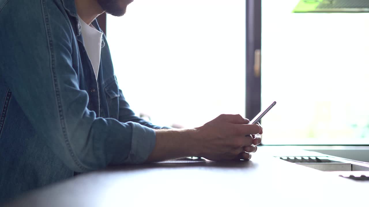 一名微笑的男子站在窗边使用智能手机。英俊的人在家里与现代小玩意共度闲暇时光视频素材