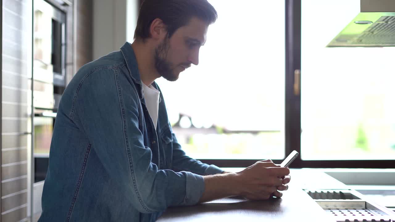 一名微笑的男子站在窗边使用智能手机。英俊的人在家里与现代小玩意共度闲暇时光视频素材