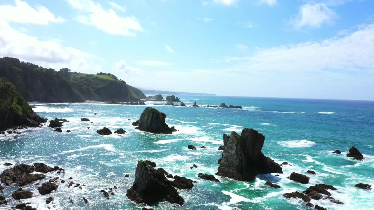 在西班牙阿斯图里亚斯的寂静海滩，蓝色的海浪拍打着岩石和崎岖的海岸线。——空中回落视频下载