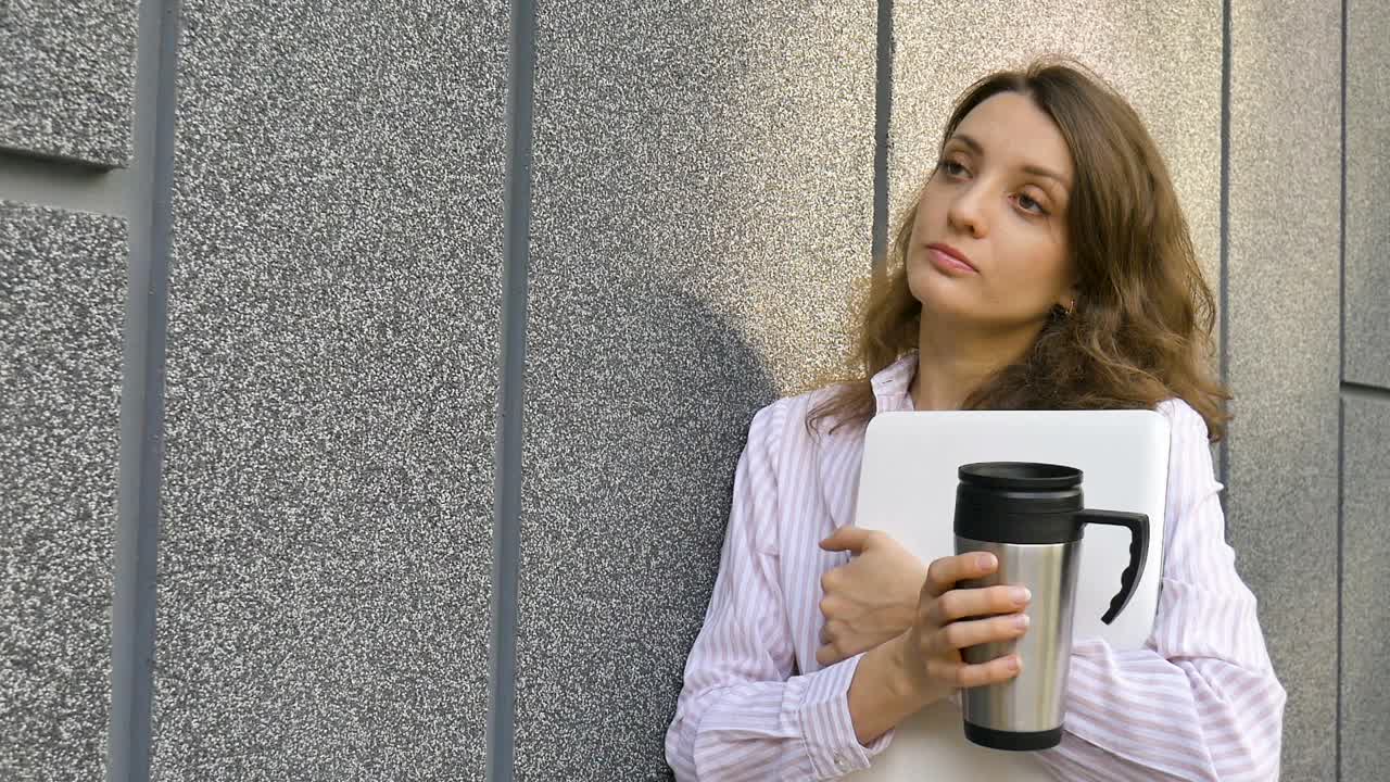 女性的肖像年轻的女人与银色笔记本电脑和一杯咖啡等待会议在深灰色的墙壁在早晨，远程工作，咖啡休息，聪明的学生概念视频素材