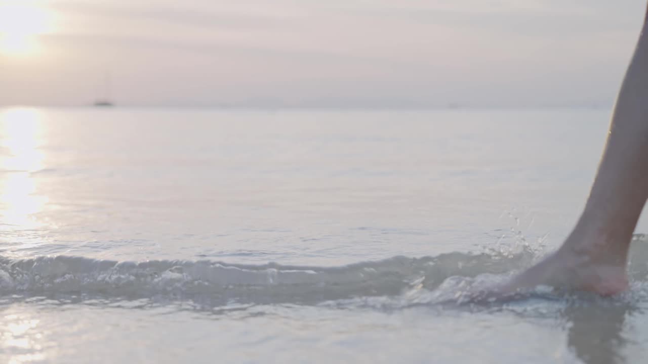 侧视年轻女子的腿和脚沿着海滩上的海水波浪行走，在金色的日落时分，平静放松宁静思考，岛屿天堂静修，低角度拍摄视频素材