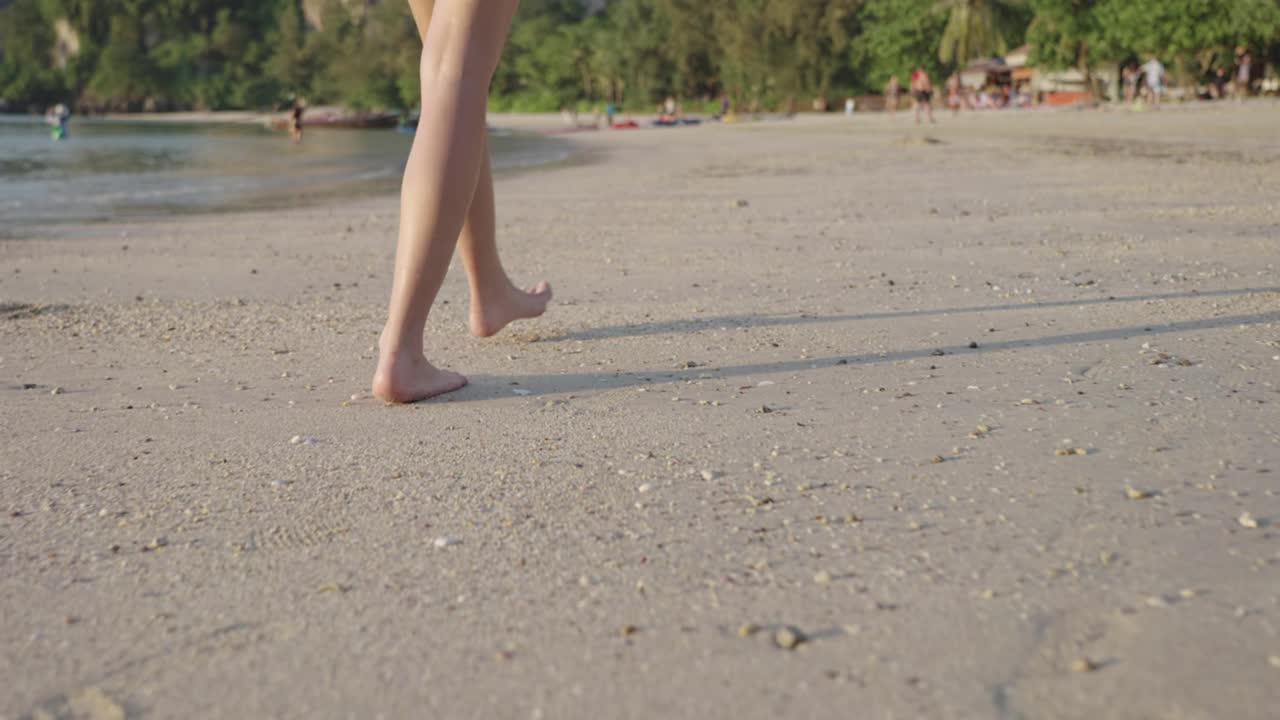 女腿赤脚沿着沙滩低潮平静海浪，后景美女悠闲漫步，夏日热带岛屿海滩，轻松漫步，下半身旅游预订度假胜地视频素材