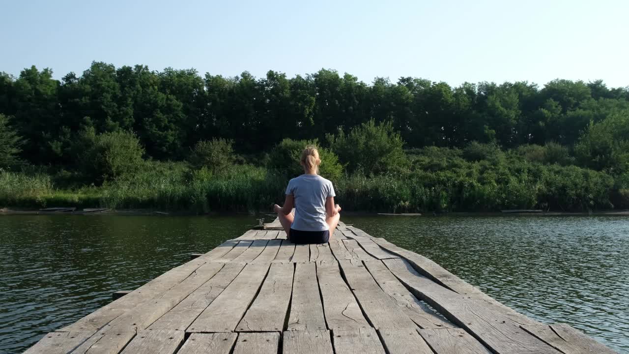 清晨，一名年轻女子坐在河上的木桥上练习瑜伽。女性以莲花坐姿冥想。自然、健康、个人活动的概念视频素材