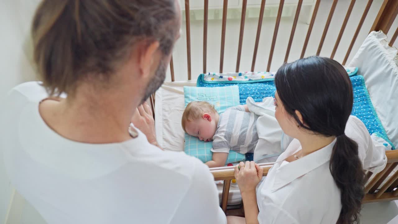 白种父母在家照顾睡在卧室里的小男孩。幸福的家庭，美丽的有爱心的妈妈和爸爸把毯子盖在舒适的睡午觉的小婴儿在床上。视频素材